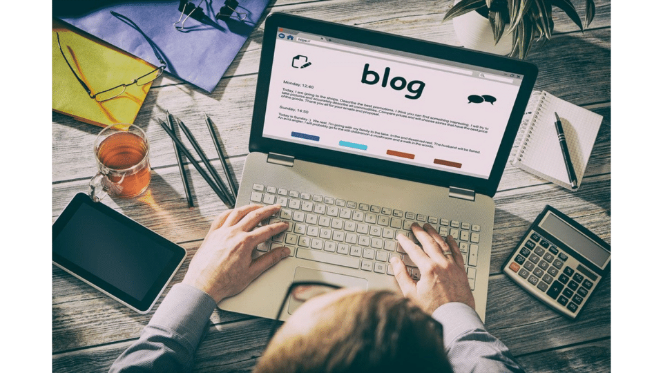 Overhead view of a person typing on a laptop with a blog page open on the screen. The workspace includes a tablet, a glass of tea, several pens, a calculator, and notepads on a wooden desk.